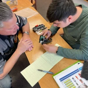 Two people are seated at a table with notes and various tools. The younger person is showing the older person a small electronic device. They are engaged in a discussion or examination.
